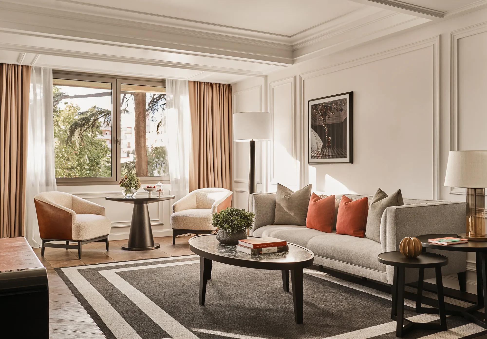 Suite living room with a grey sofa and orange cushions, two armchairs around a small table, and a window onto trees.