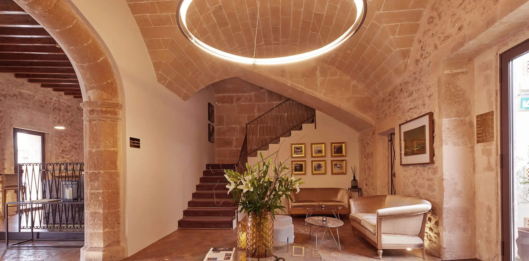 Lobby at Sa Creu Nova with a vaulted brick ceiling lit by a circular ring light, a stone archway, a staircase and cream sofas beside a gallery of framed art