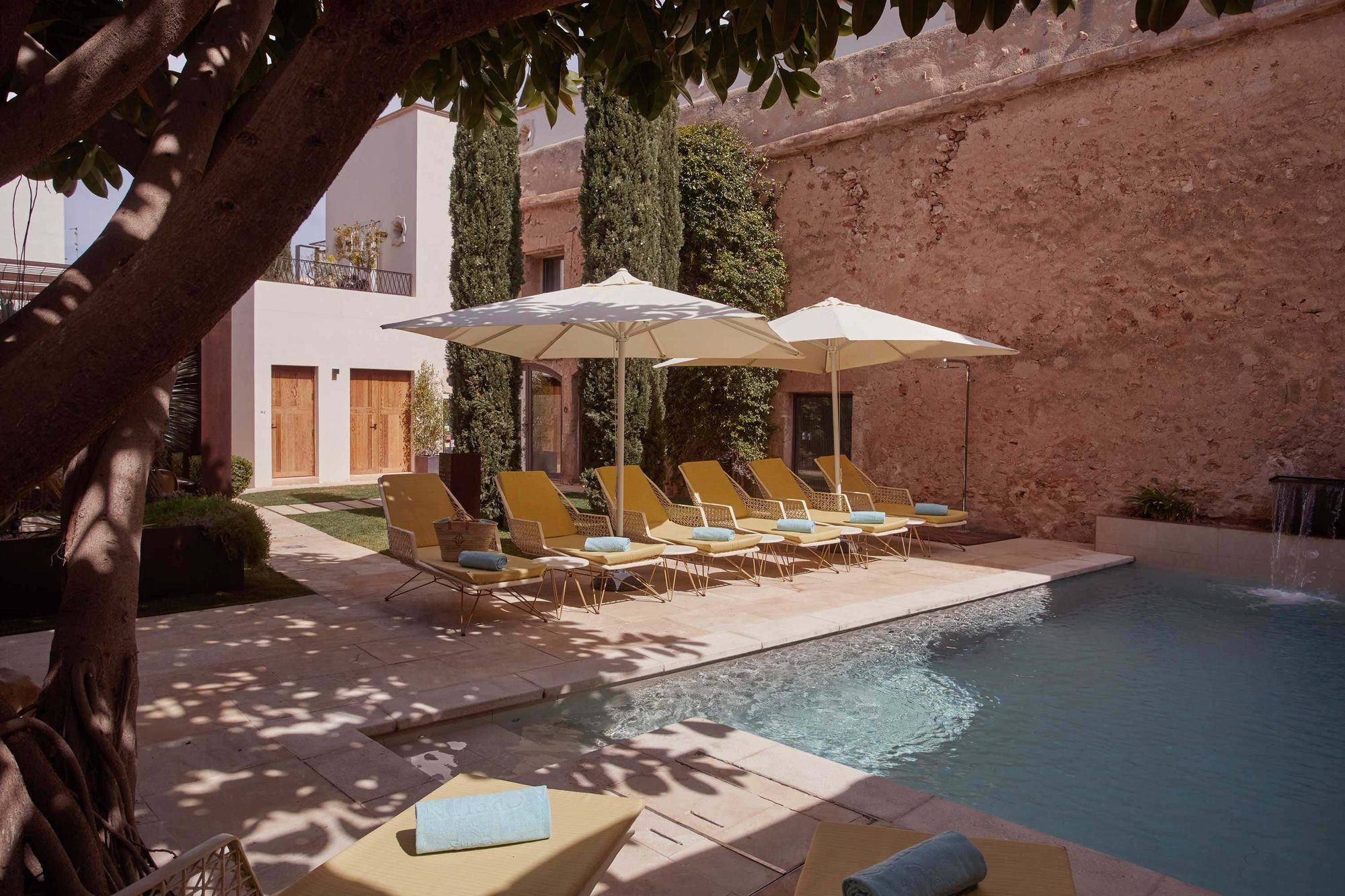 Courtyard pool at Sa Creu Nova bordered by an old stone wall and cypress trees, with mustard-yellow loungers and umbrellas along the tiled deck