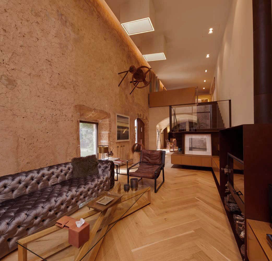Two-level Penthouse living room at Sa Creu Nova with a raw stone wall, a tufted leather chesterfield sofa and herringbone parquet flooring beneath a mezzanine loft
