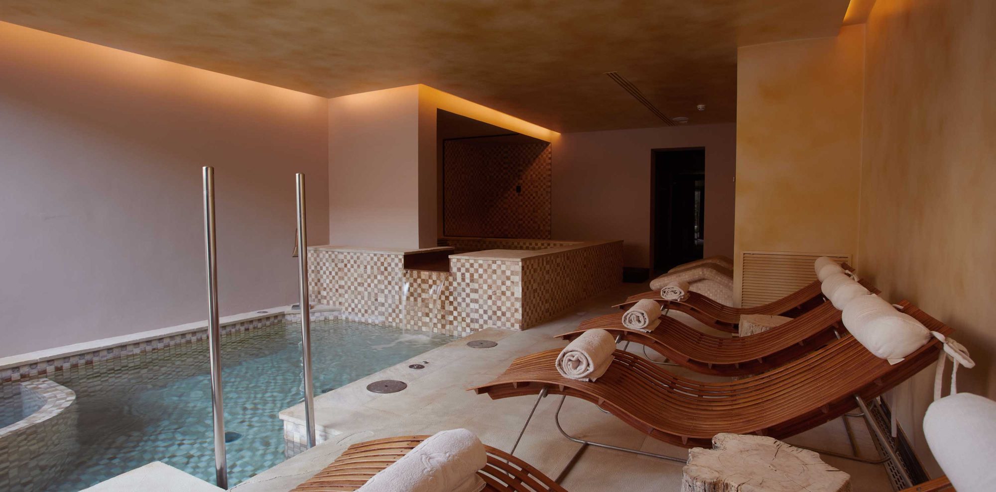 Indoor spa pool at Sa Creu Nova with a mosaic tile edge and curved wood loungers with rolled towels under warm ambient lighting