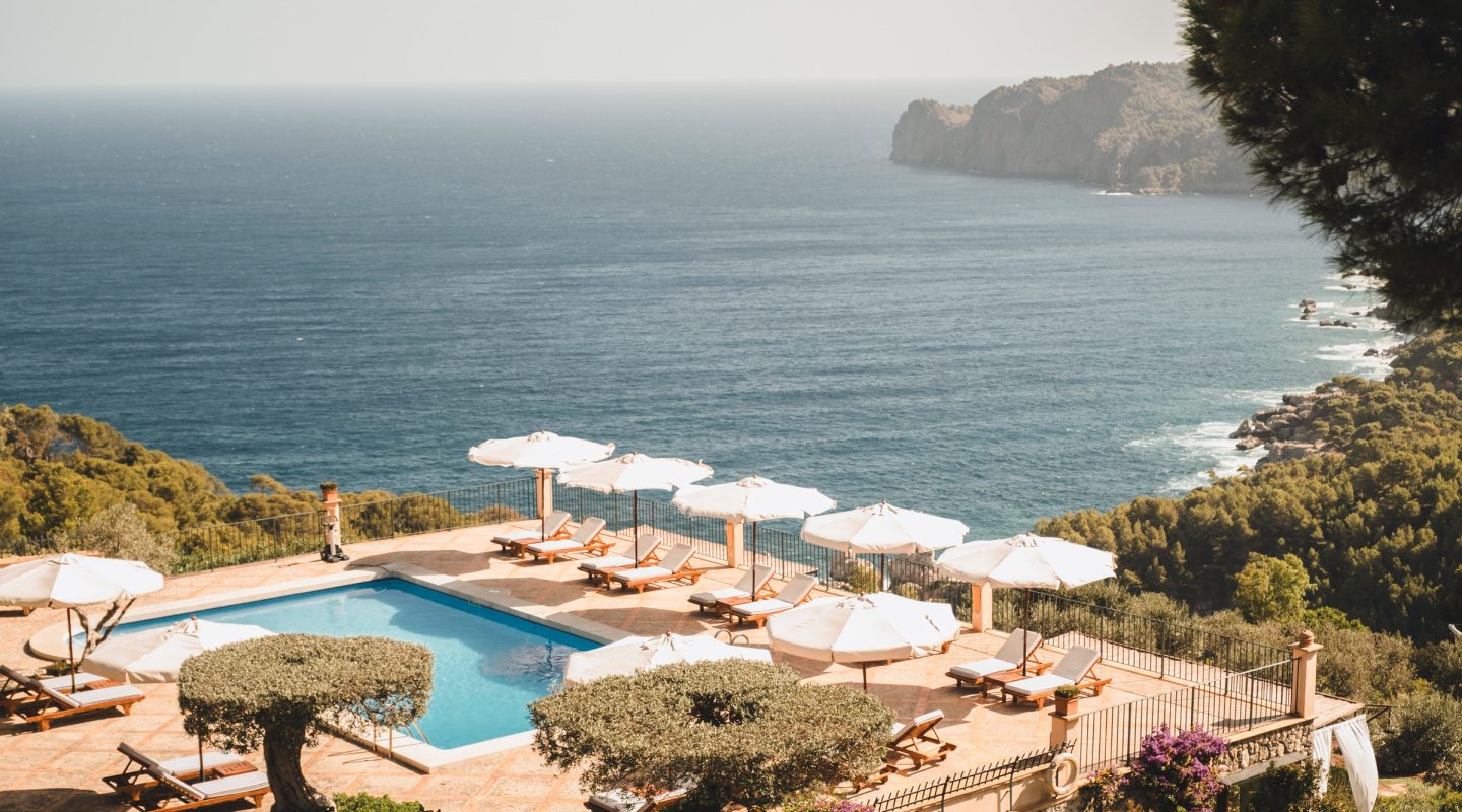Cliffside pool at Sa Pedrissa with white umbrellas and sunbeds on a terracotta tile deck overlooking the Mediterranean and a rocky headland