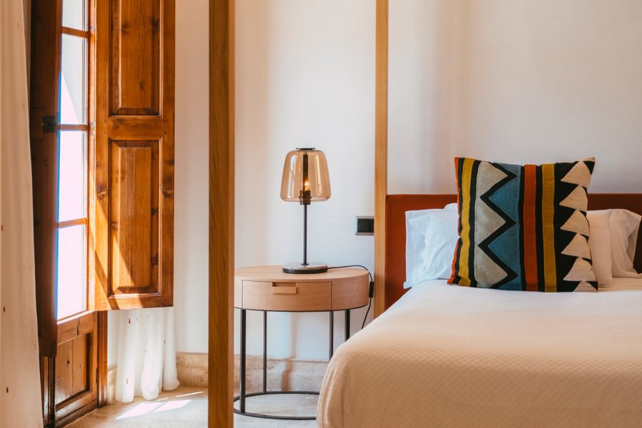 Suite Jardin at Sa Pedrissa with a white bed under a wood four-poster frame, a geometric-patterned throw pillow and a round nightstand with an amber glass lamp