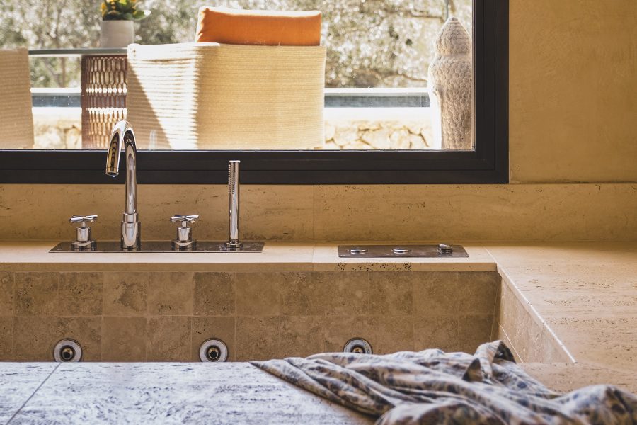 Tiled bathtub at Sa Pedrissa with chrome fixtures beneath a window looking onto a terrace with a woven basket and orange cushion