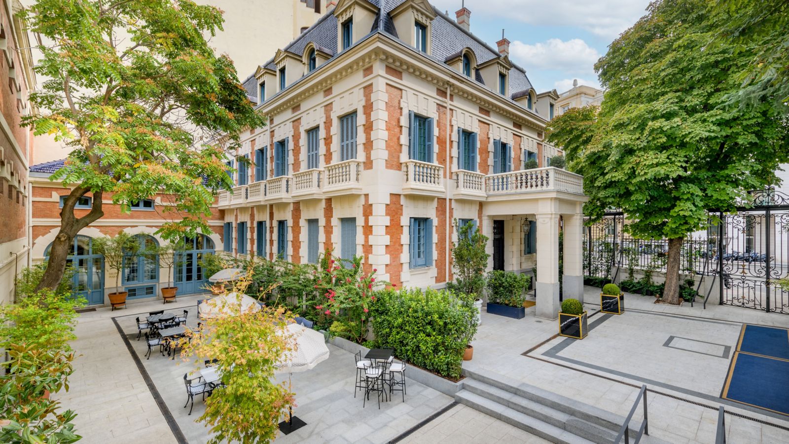 The brick-and-stone mansion with blue shutters and a mansard roof seen across its paved courtyard, where cafe tables, parasols and planted greenery fill the terrace.
