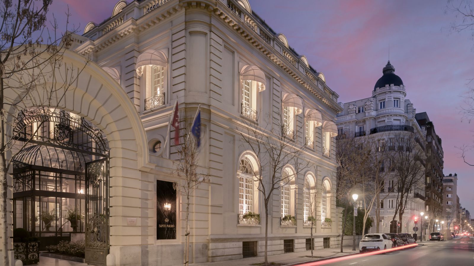 The palace's street facade at dusk: a white neoclassical corner with lit arched windows, an arched iron gate with a glass canopy behind it, and a black marble portal lettered Santo Mauro.