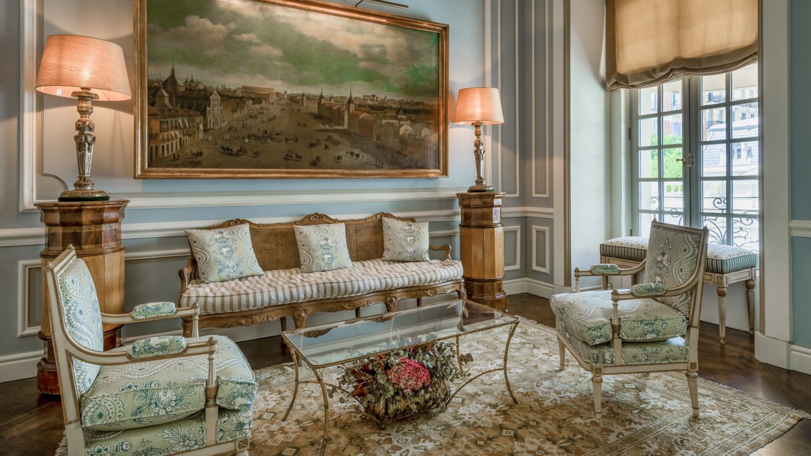 A salon with a caned antique settee and two floral armchairs around a glass table, flanked by column lamps beneath a large gilt-framed cityscape painting.