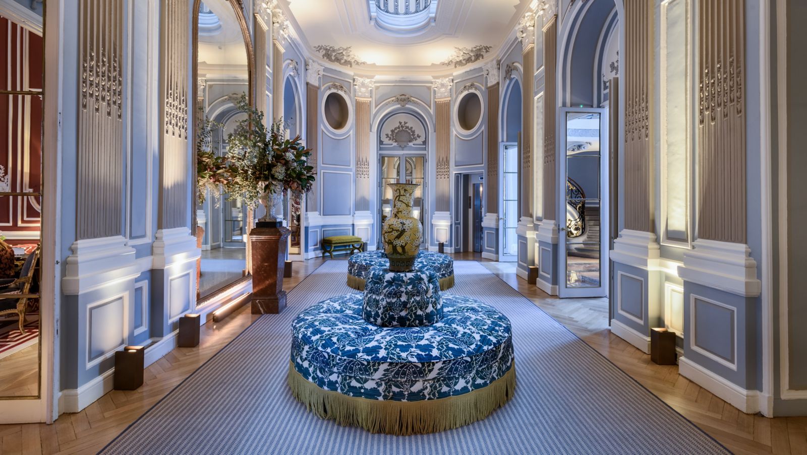 An oval hall with pale blue panelled walls, arched mirrored doorways, a domed skylight and two round blue-and-white upholstered ottomans under a large vase.