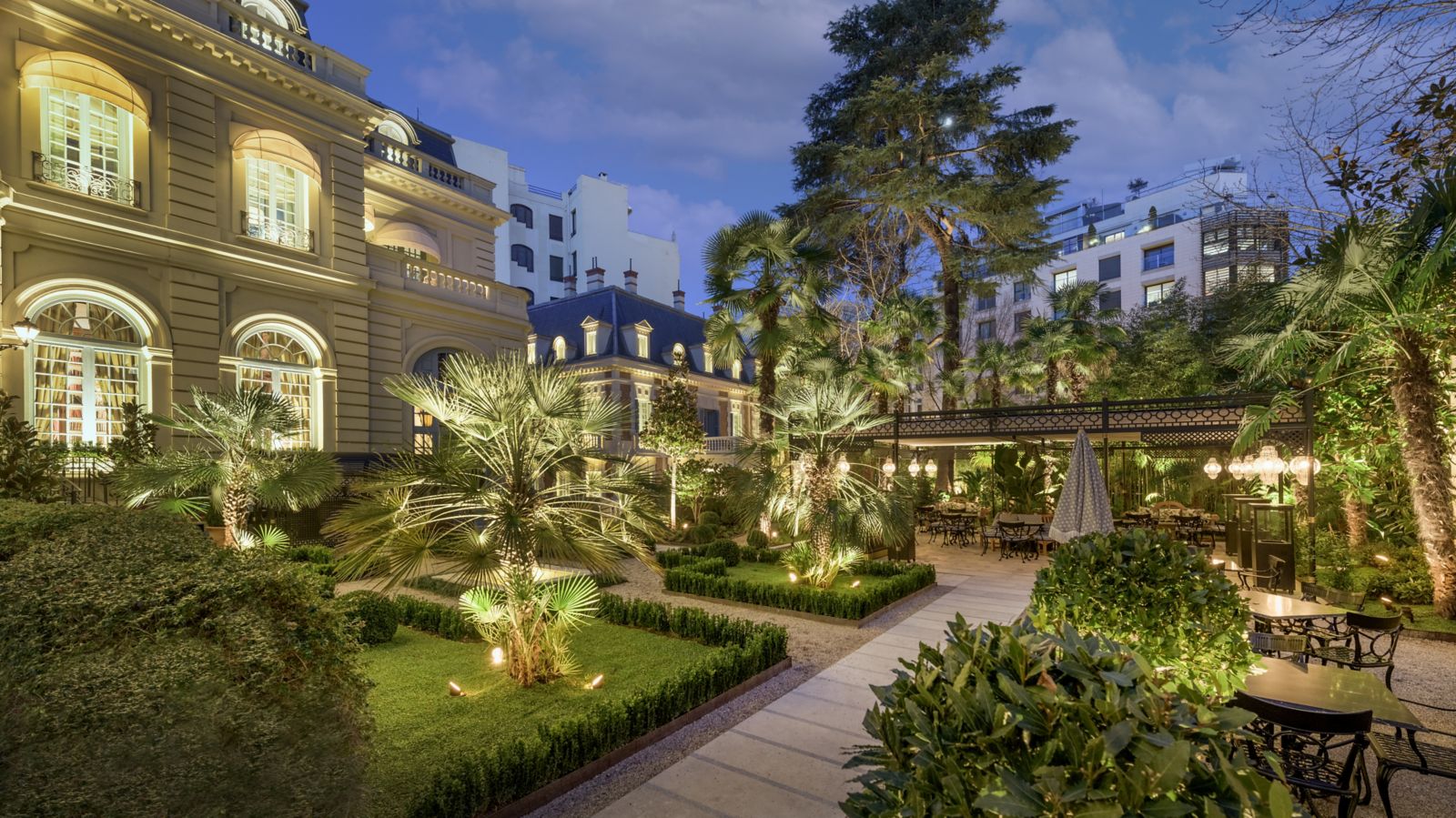 The garden at dusk: lit palms and clipped box parterres along gravel-edged paths, with a covered dining terrace of lantern-lit tables at the far end.