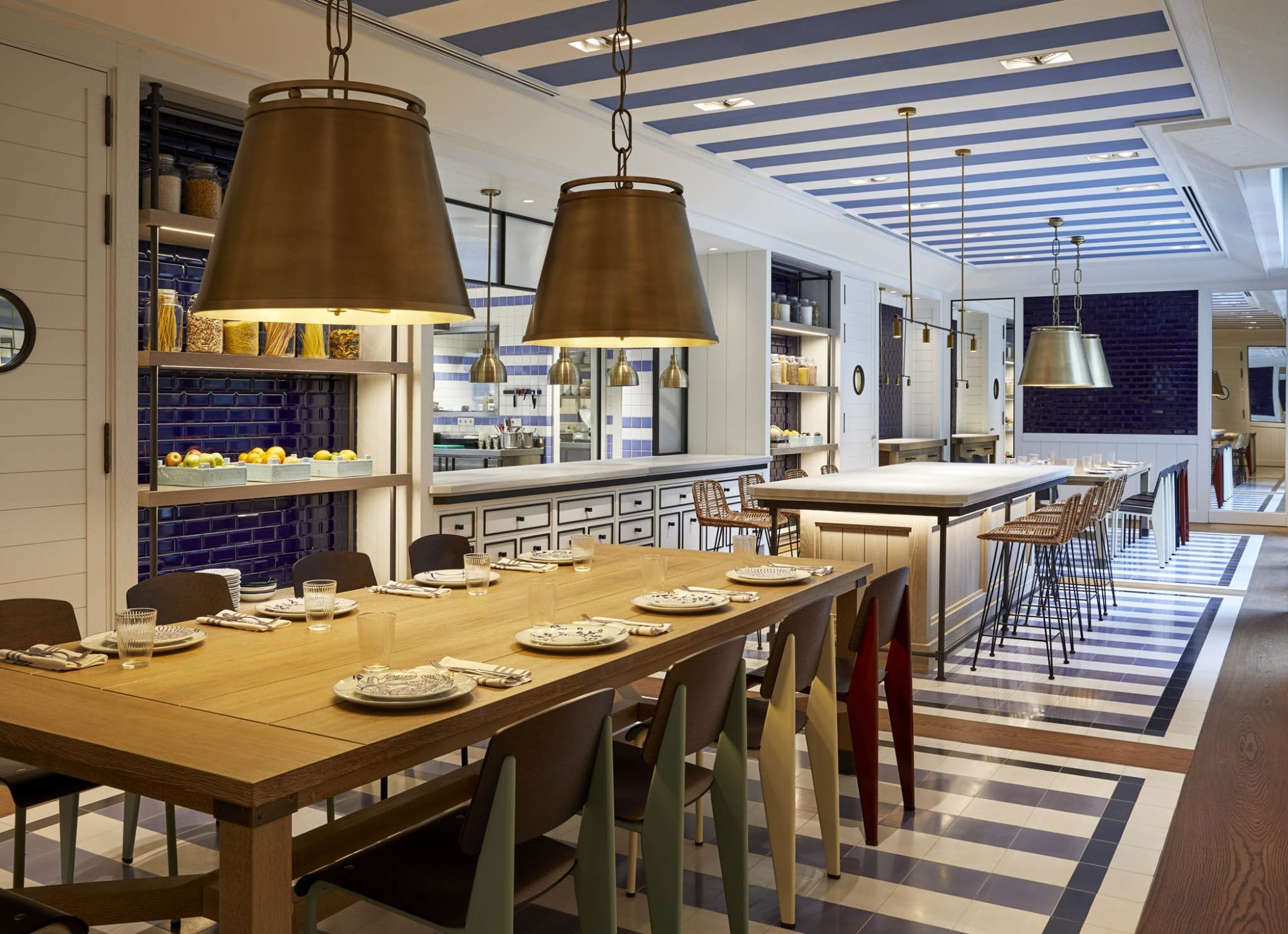 Communal dining table at Seventy Barcelona's restaurant set beneath brass pendant lamps, with an open kitchen behind and a blue-and-white striped tile floor