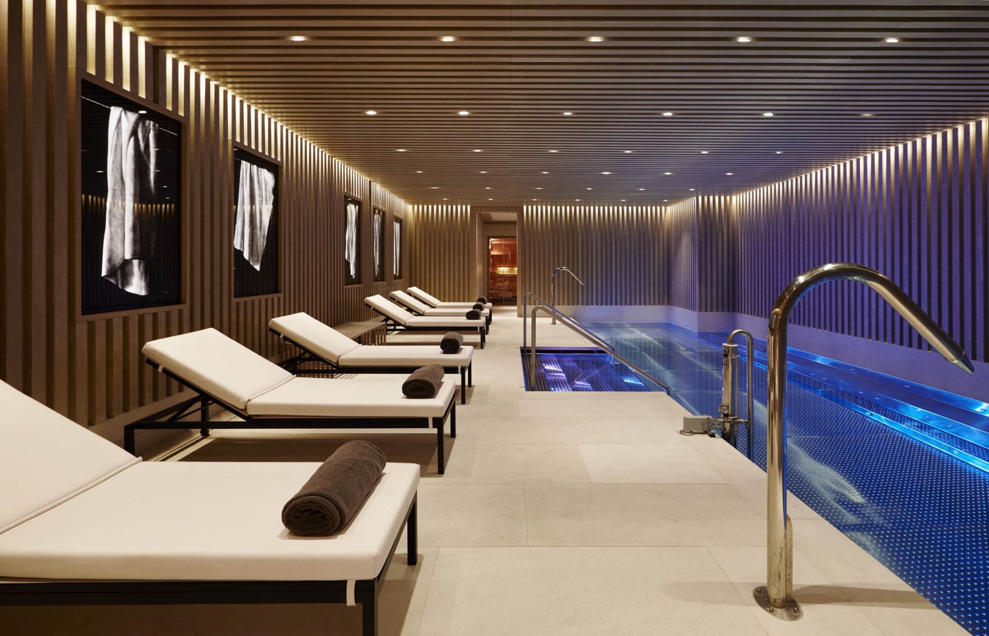 Indoor pool at Seventy Barcelona with a slatted wood ceiling, white loungers with rolled towels along one side and blue-lit water