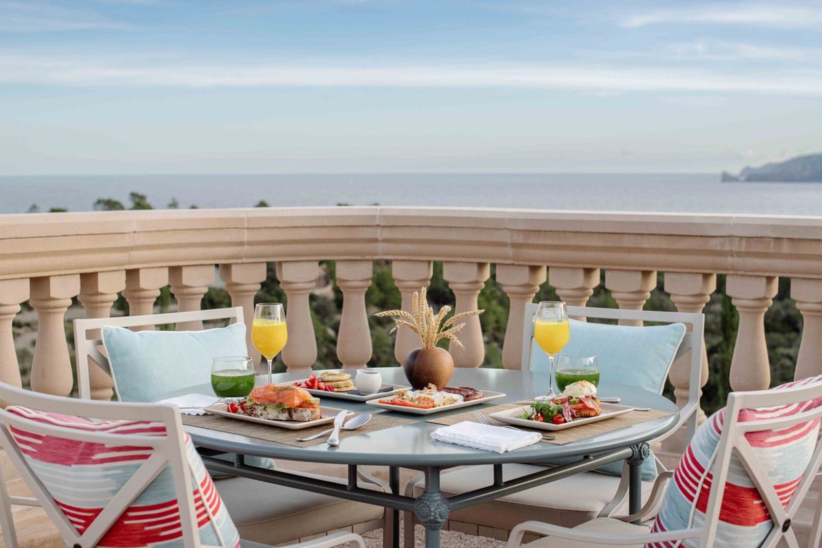 Breakfast set on a curved stone balcony at Son Bunyola with striped chairs, orange juice and plated food overlooking the sea