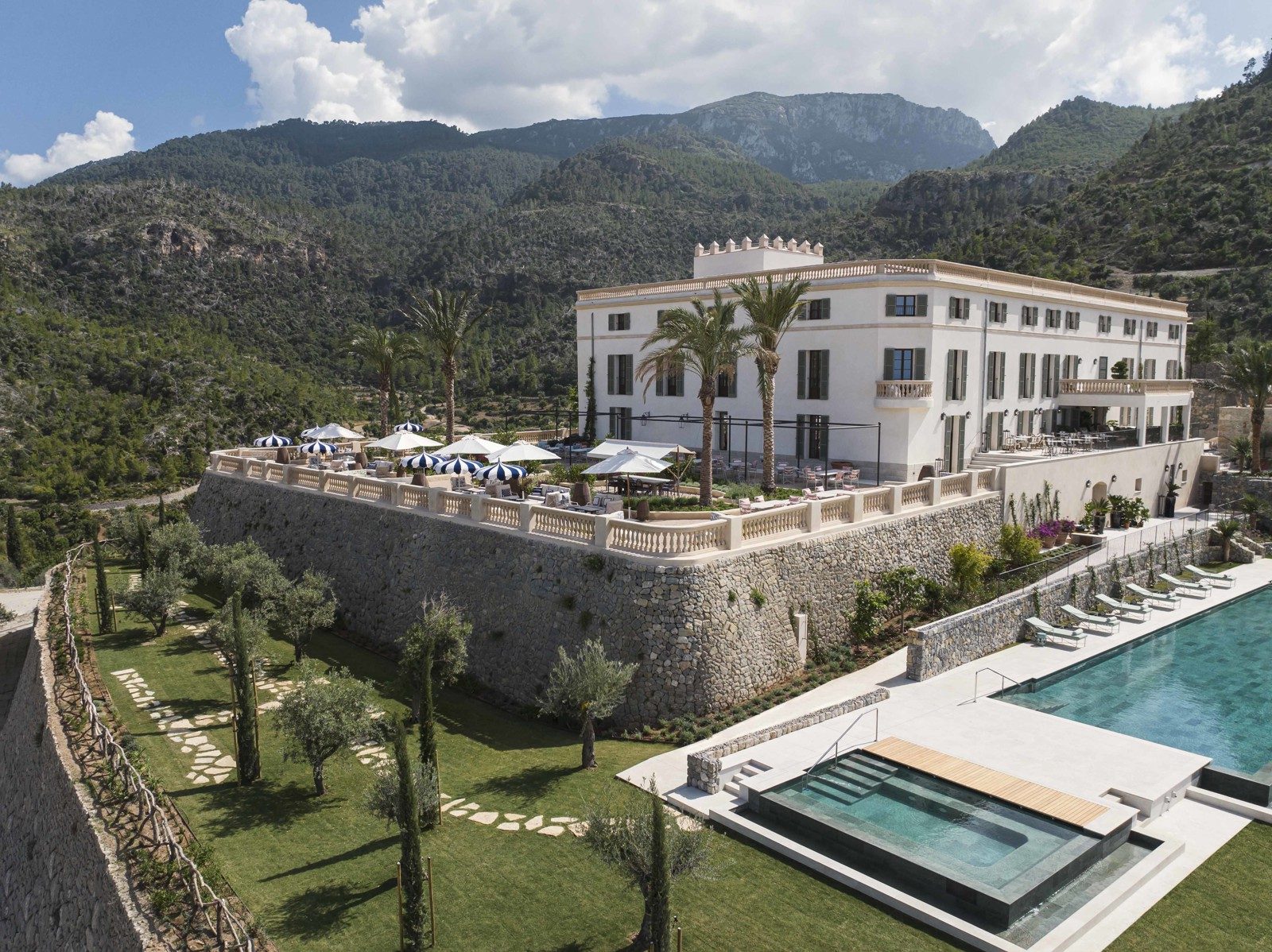Aerial view of Son Bunyola's historic manor house with a crenellated tower, terraced stone gardens and a pool below, set against the Tramuntana mountains