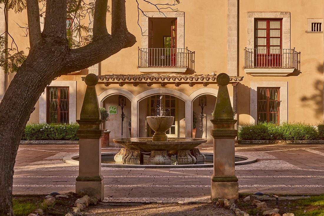 Ochre-coloured manor house facade at Son Julia with red-shuttered French balconies and a stone fountain in the courtyard below