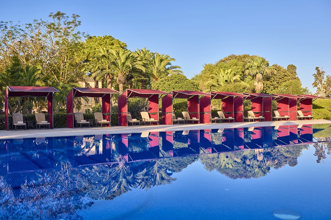 Pool at Son Julia lined with red canopy cabanas and loungers, palm trees reflected in the still water