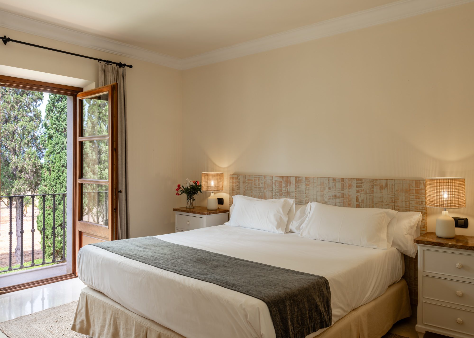 Guest room at Son Julia with a white bed and grey throw, a textured woven headboard, and a French balcony door open to a garden of cypress trees