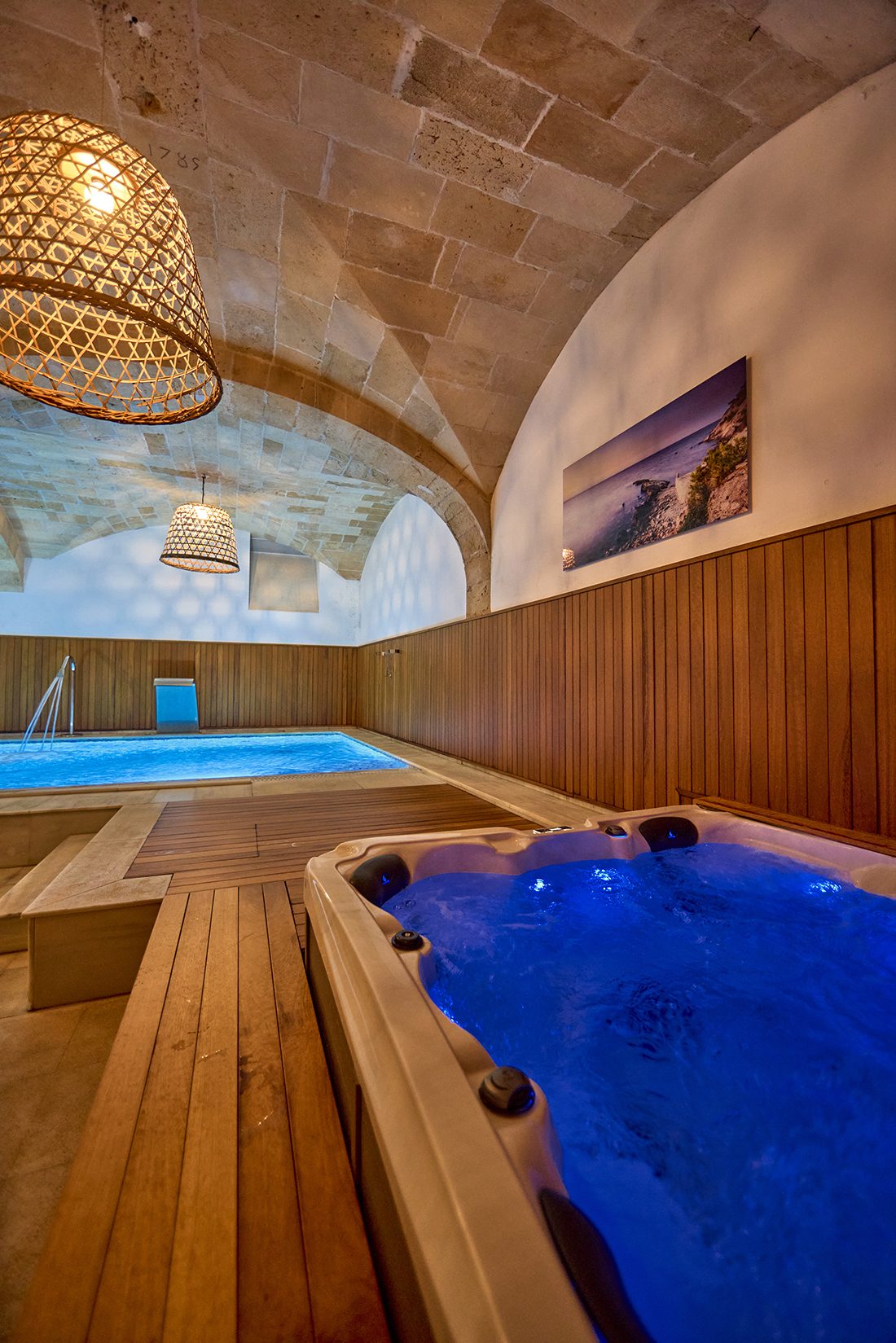 Indoor spa at Son Julia with a vaulted stone ceiling, woven rattan pendant lamps, an illuminated hydromassage tub and a small indoor pool beyond