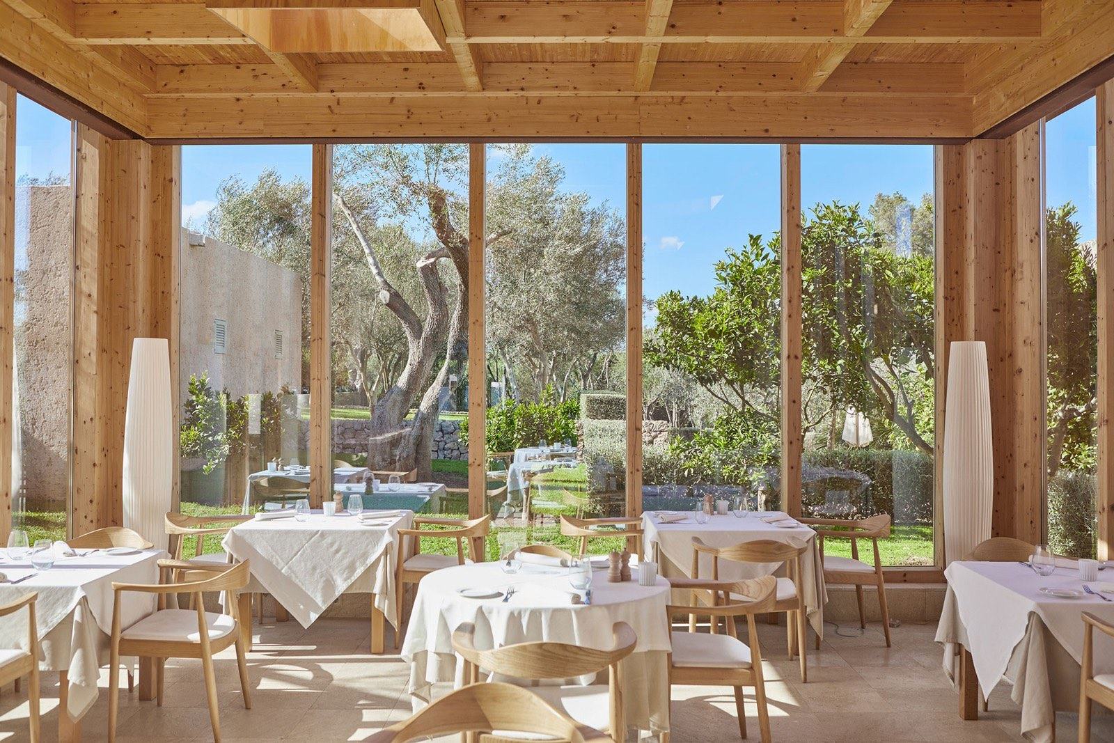 Dining room with tables laid in white linen, wooden ceiling beams and glass walls onto olive trees