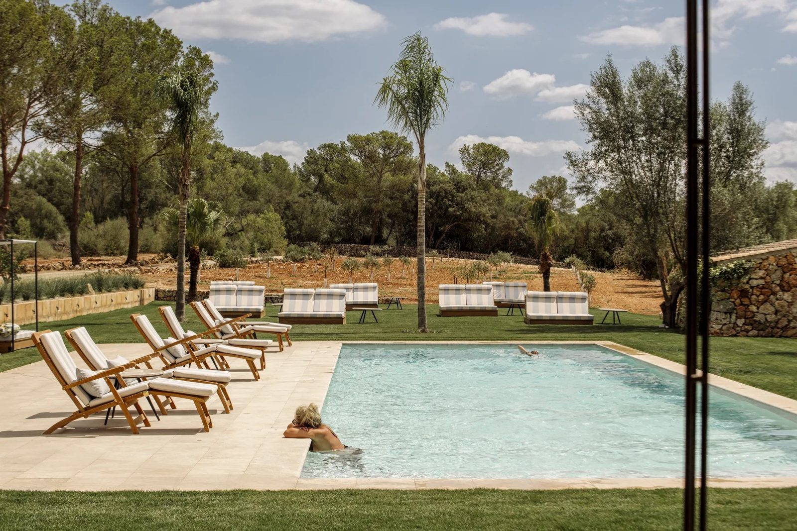 Outdoor pool with striped sun loungers and daybeds, palms and olive trees behind