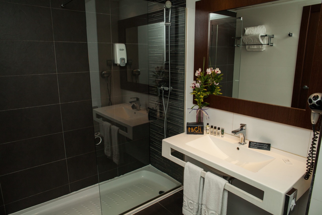 A hotel bathroom with a glass-walled shower, a white countertop sink with toiletries and flowers, and a täCH-branded towel folded on the rail.