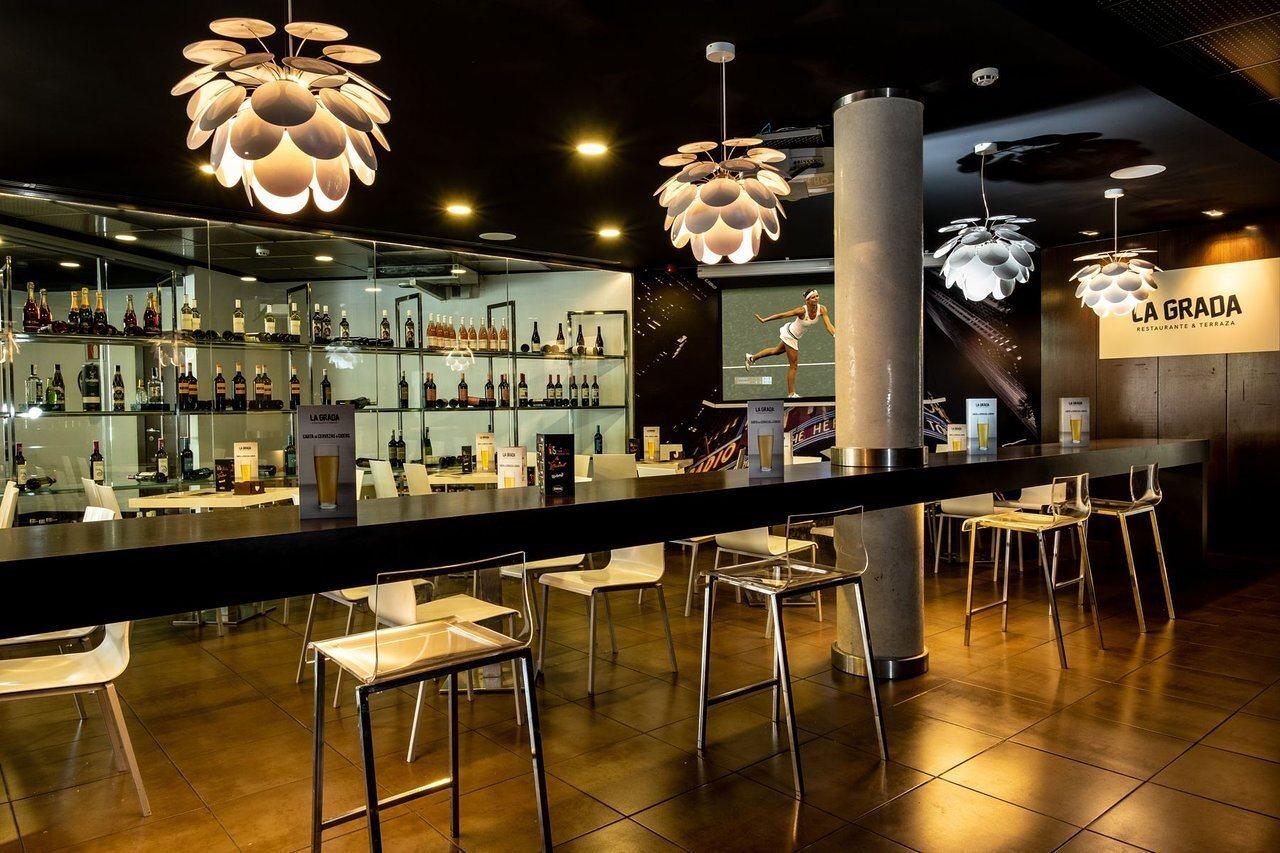 The La Grada bar and restaurant, a long bar counter with high stools, shelves of bottles, hanging pendant lights, and a lit "LA GRADA RESTAURANTE & TERRAZA" sign on the wall.