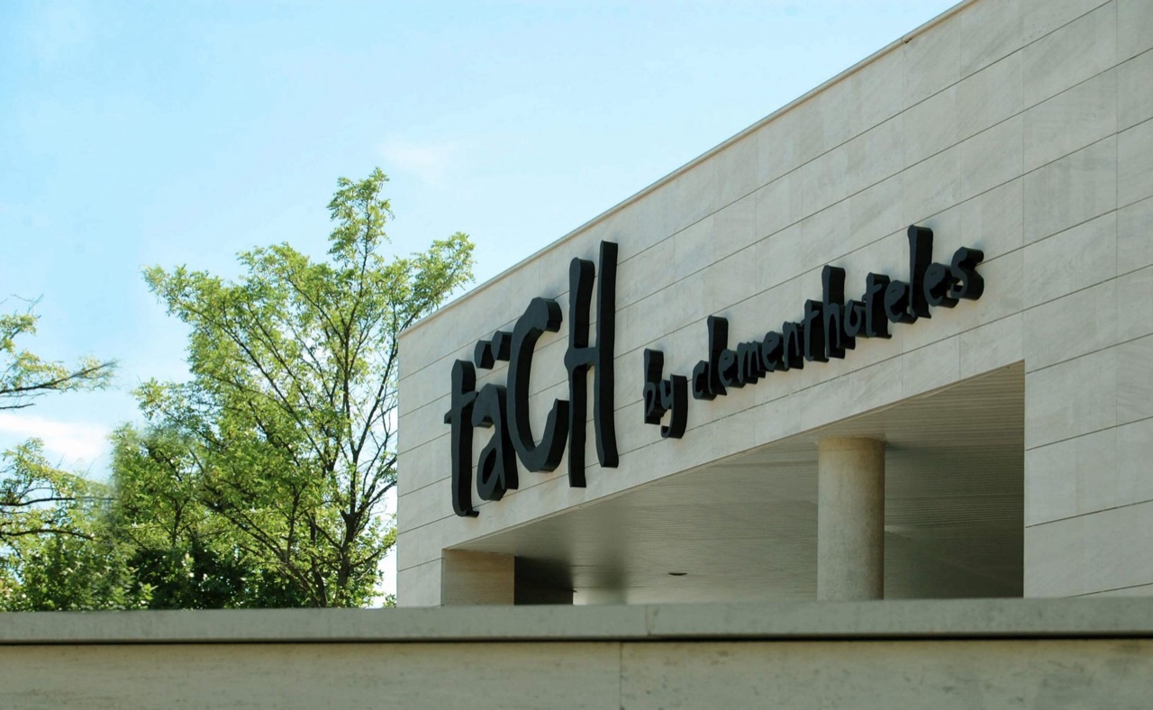 A close-up of the building's facade against a blue sky, with the raised black lettering "täCH by clementhoteles" and a tree in the foreground.