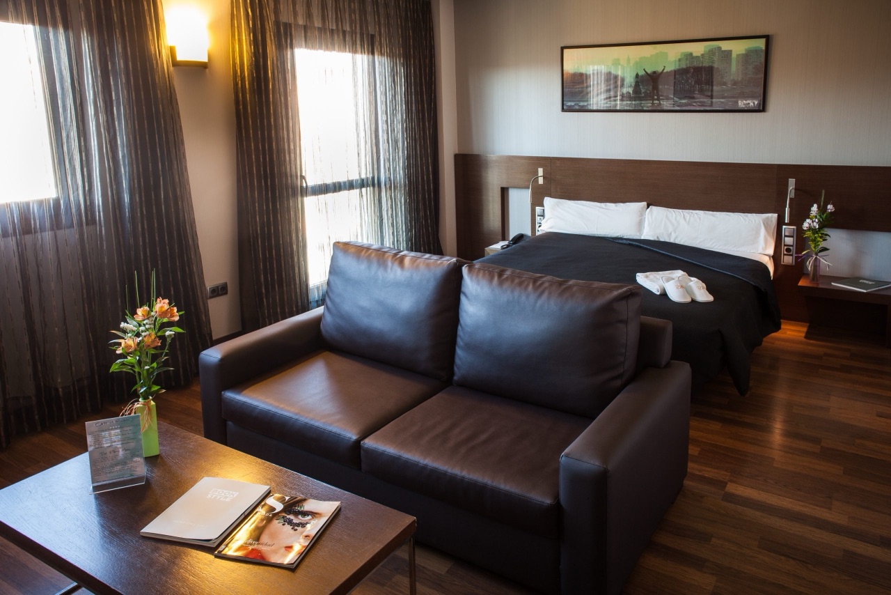A hotel room with a dark leather sofa in front of a bed with black bedding, white pillows and slippers, sheer curtains and a framed print on the wall.