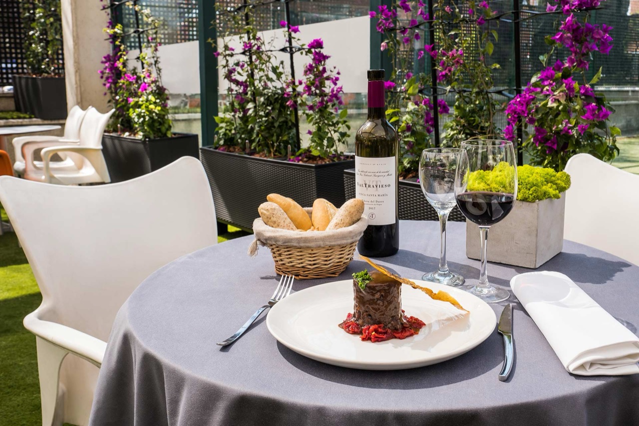 A set table on an outdoor terrace with a plated dish, a bread basket, a bottle and glasses of red wine, framed by potted flowering plants.