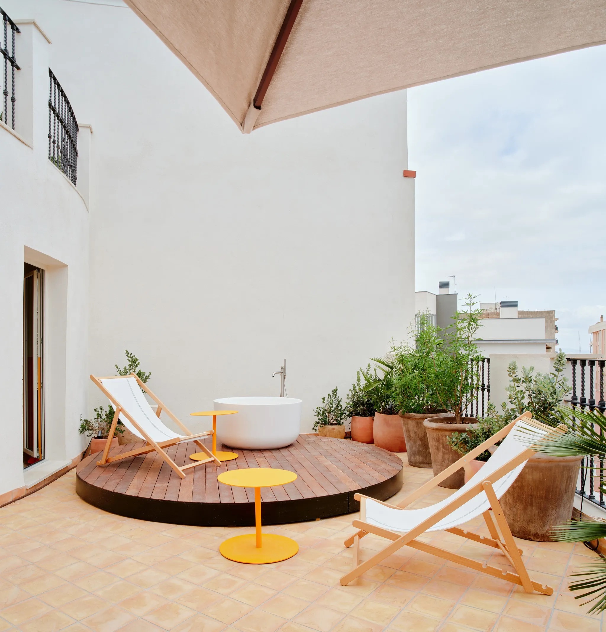Private wood-decked terrace with a freestanding soaking tub, deck chairs, and potted plants against a white facade