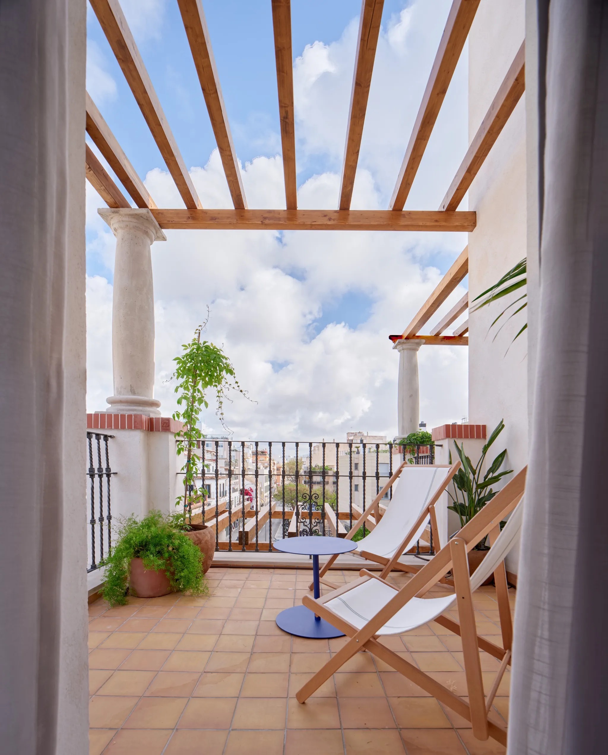 Rooftop terrace under a wood pergola with deck chairs, potted plants, and a view over Palma rooftops