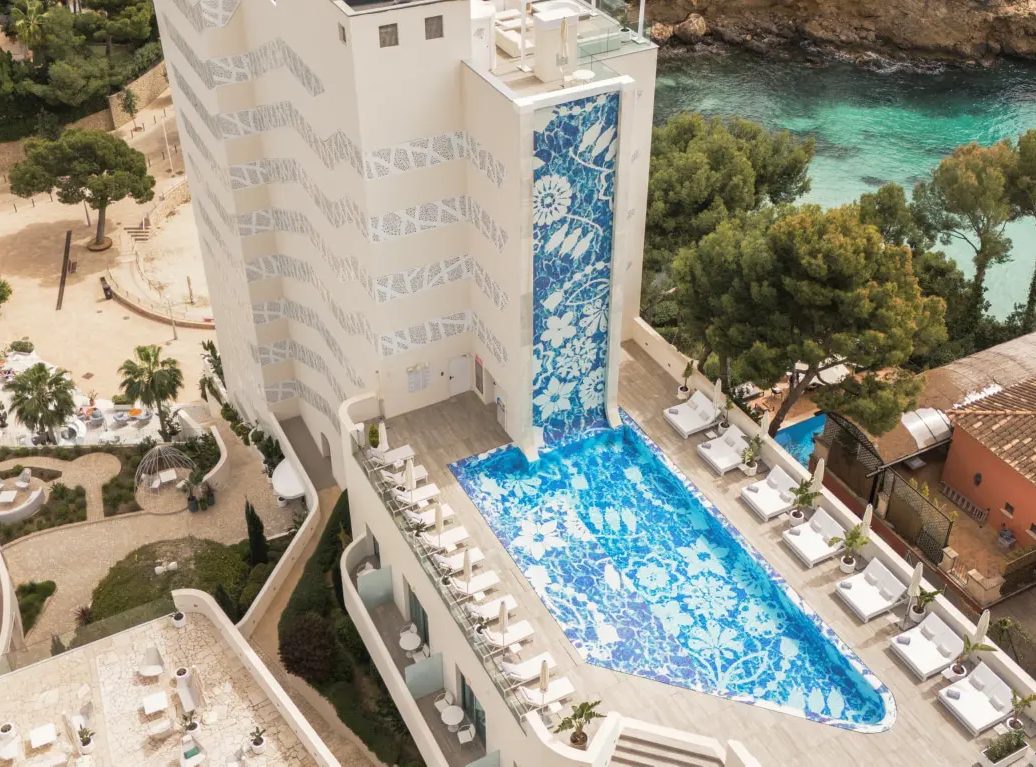 Aerial view of The Donna Portals' mosaic-tiled tower facade above a matching mosaic-patterned pool, loungers and a turquoise cove behind