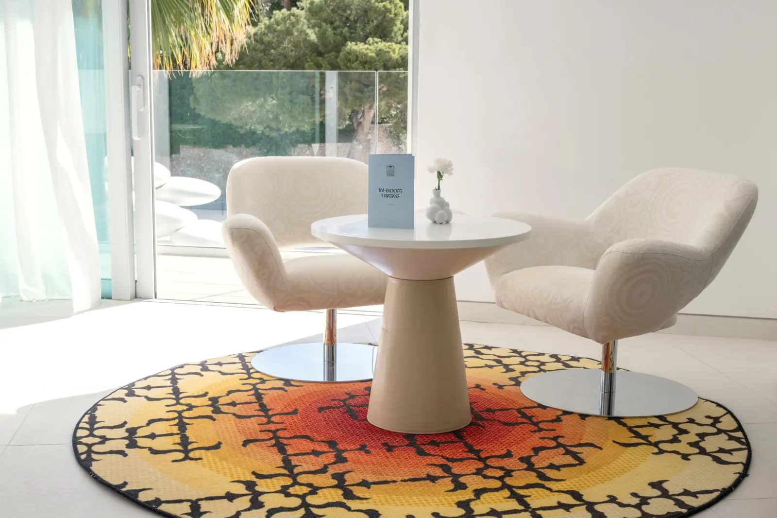 Marcel Wanders Executive Room seating area at The Donna Portals with two boucle swivel armchairs, a round pedestal table and a mandala-patterned rug beside glass doors onto the terrace