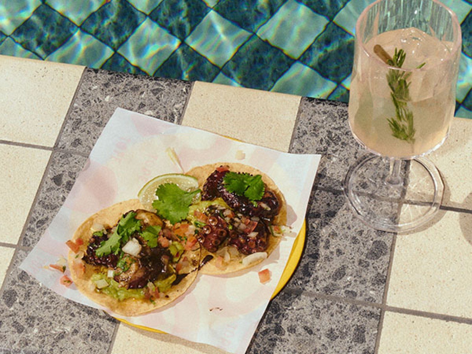 Tacos and a cocktail set on a tiled table at the edge of The Hoxton Poblenou's blue-tiled pool