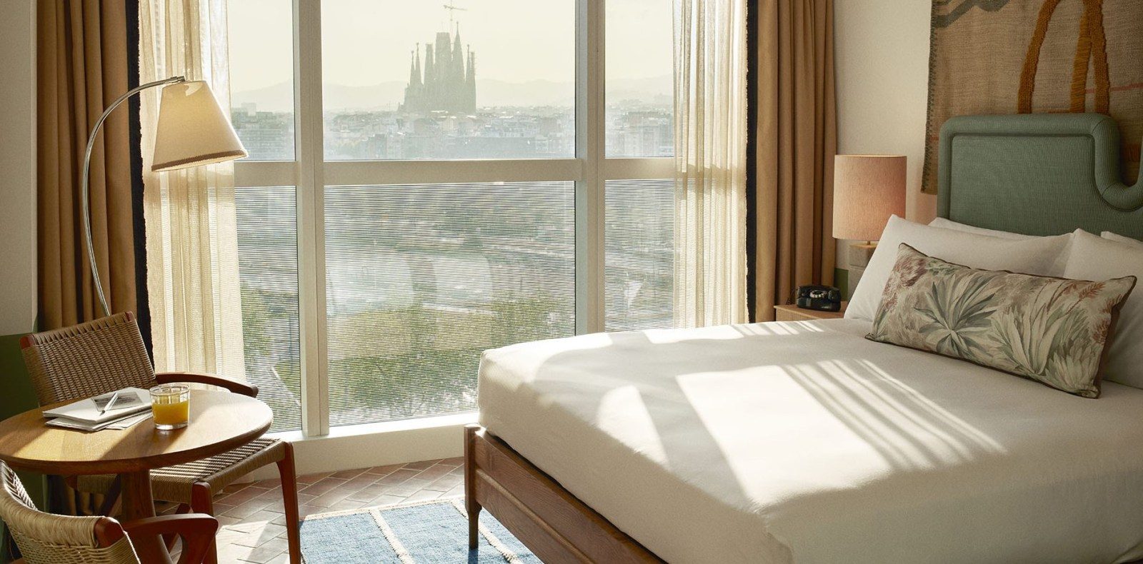 Guest room at The Hoxton Poblenou with a green upholstered headboard and floor-to-ceiling windows showing a distant view of the Sagrada Familia