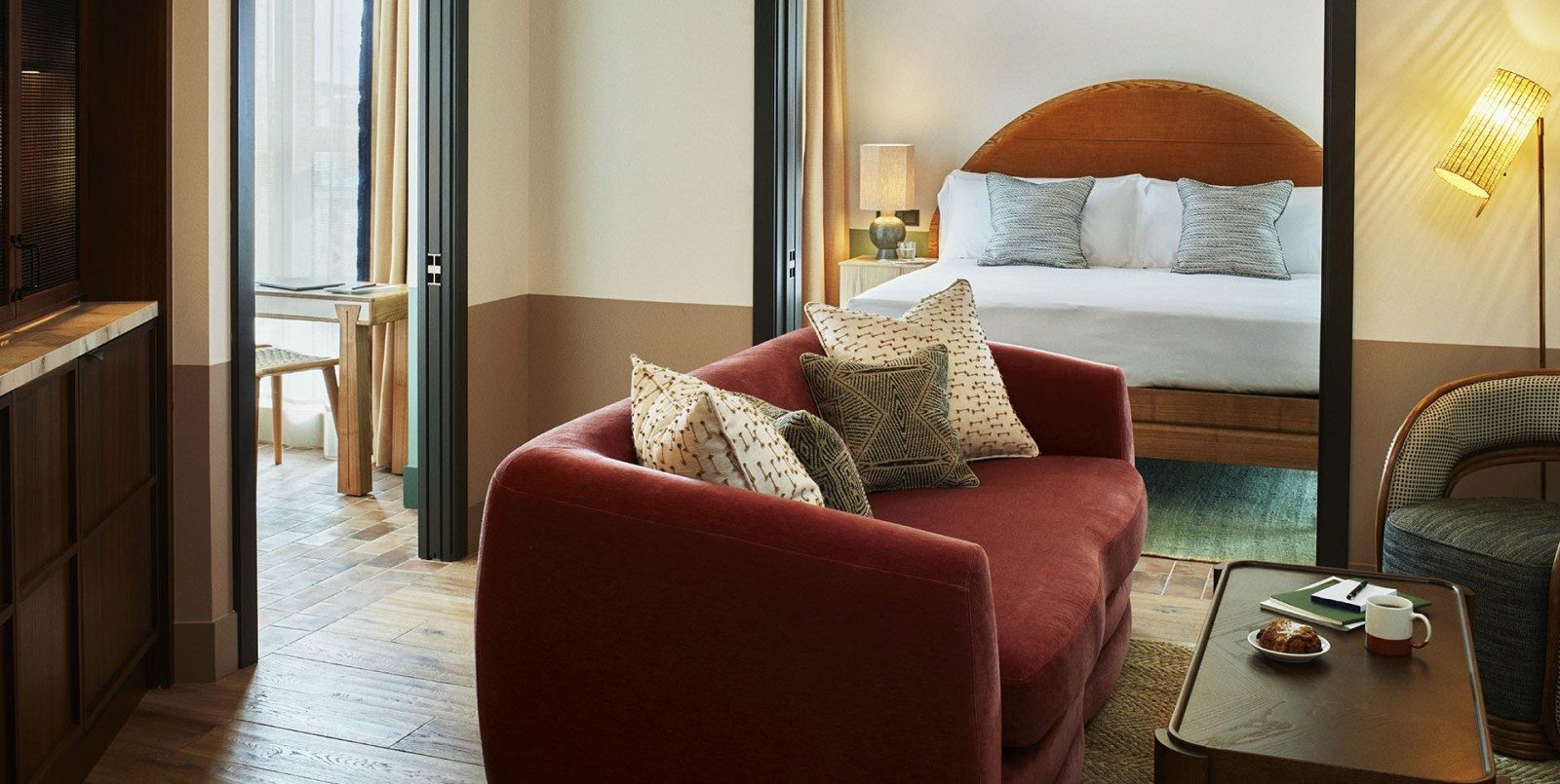 Suite at The Hoxton Poblenou with a burgundy velvet sofa holding patterned cushions, an arched wood headboard visible through open doors and a wicker armchair