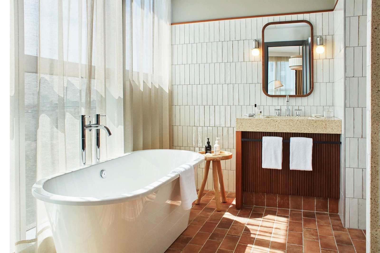 Bathroom at The Hoxton Poblenou with a freestanding tub, terracotta floor tile, white subway-style wall tile and a fluted wood vanity