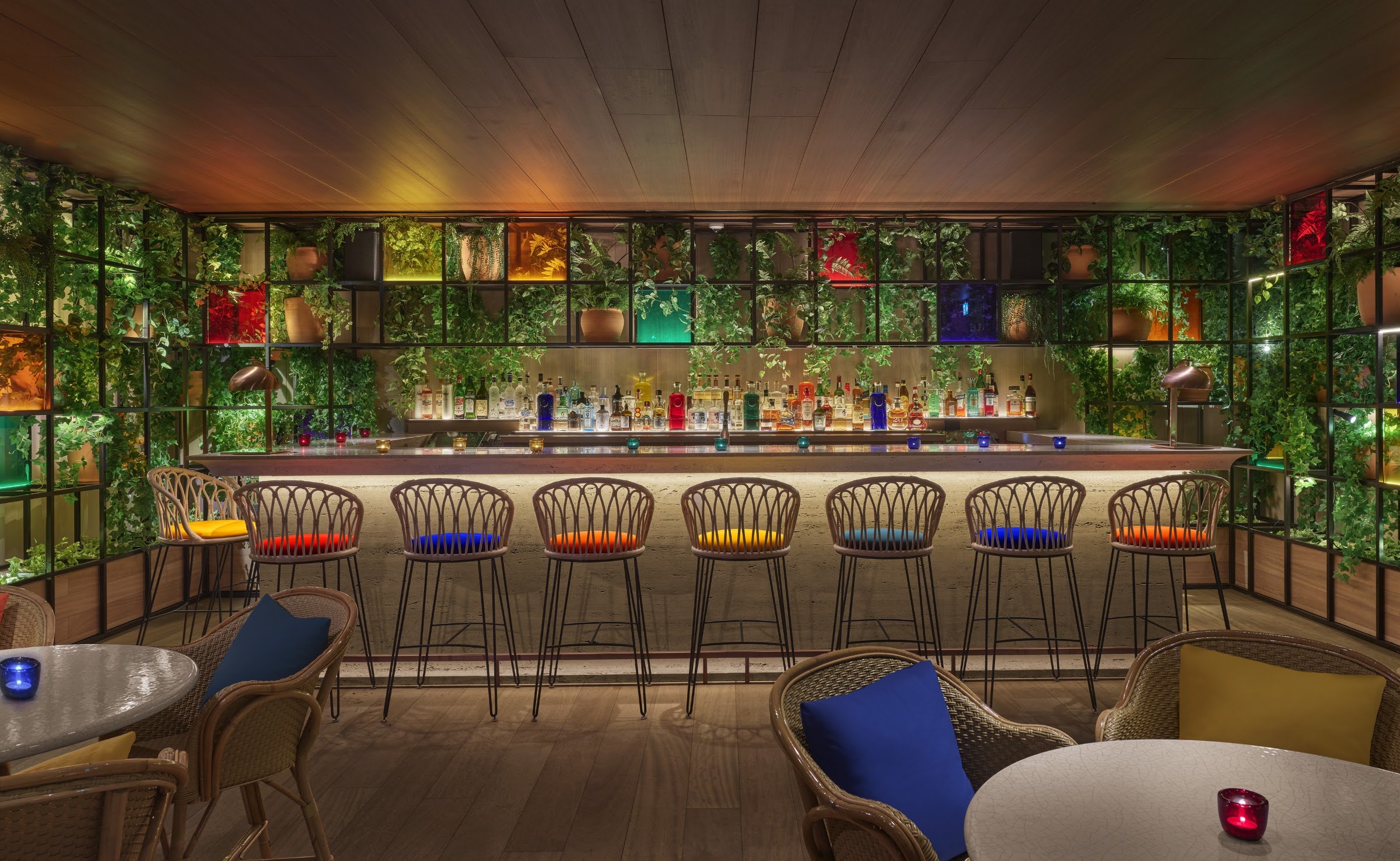 A bar with colourful bottles set into a plant-covered wall, a row of rattan stools and blue-cushioned lounge chairs.