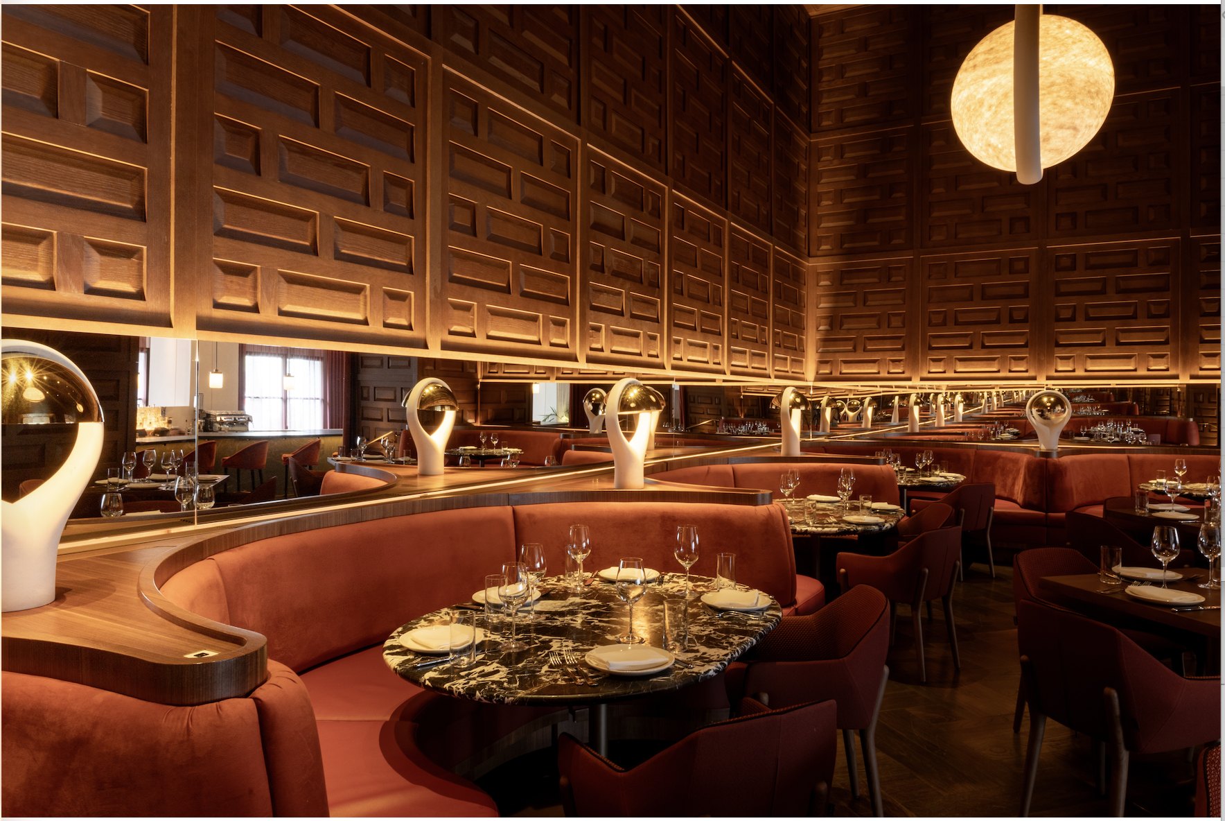 A dining room with rounded red velvet booths, marble tables and dark wood-panelled walls under a large globe pendant light.