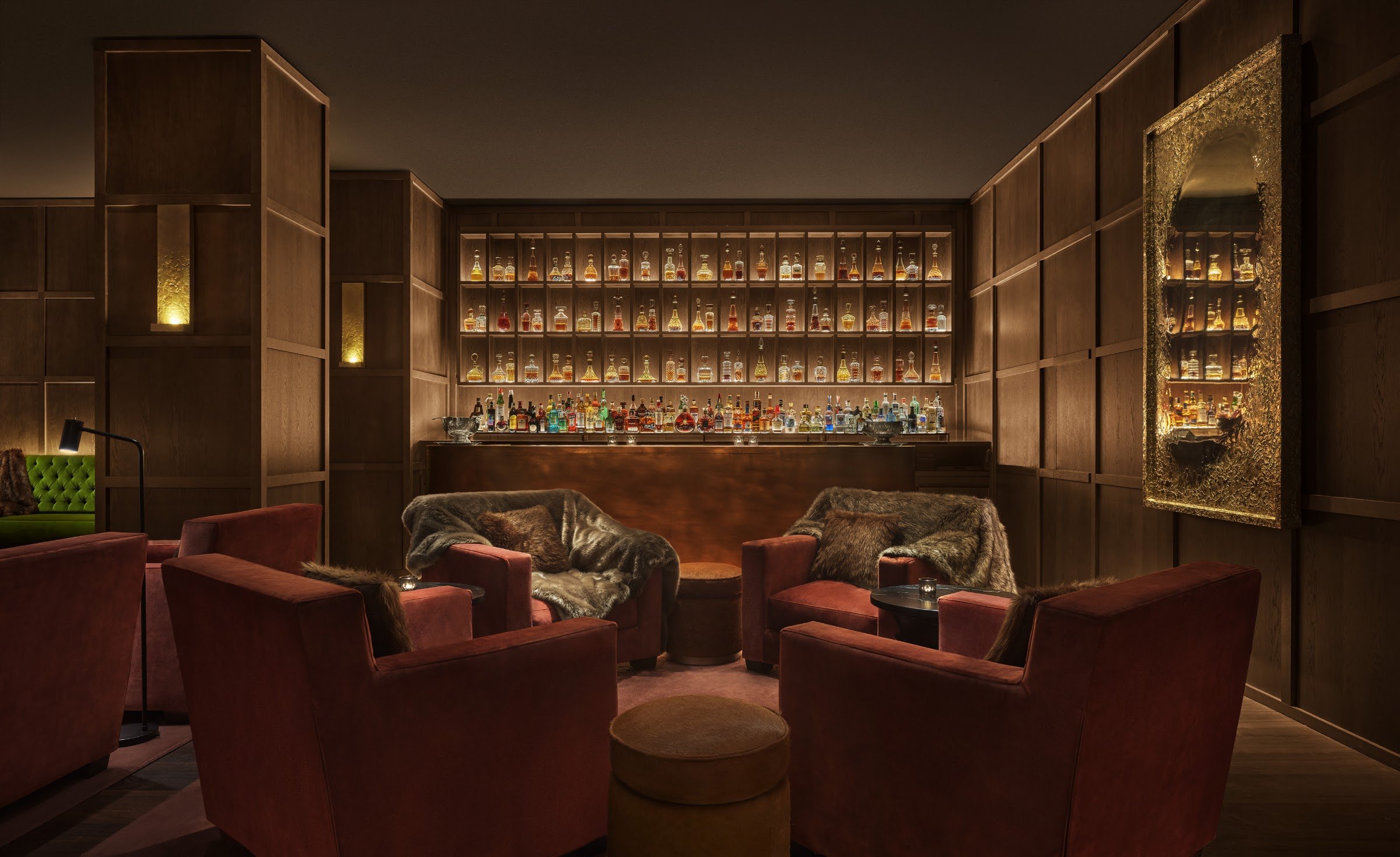 A dim lounge bar with rust-red armchairs, a low round table and a back bar of glowing shelves.
