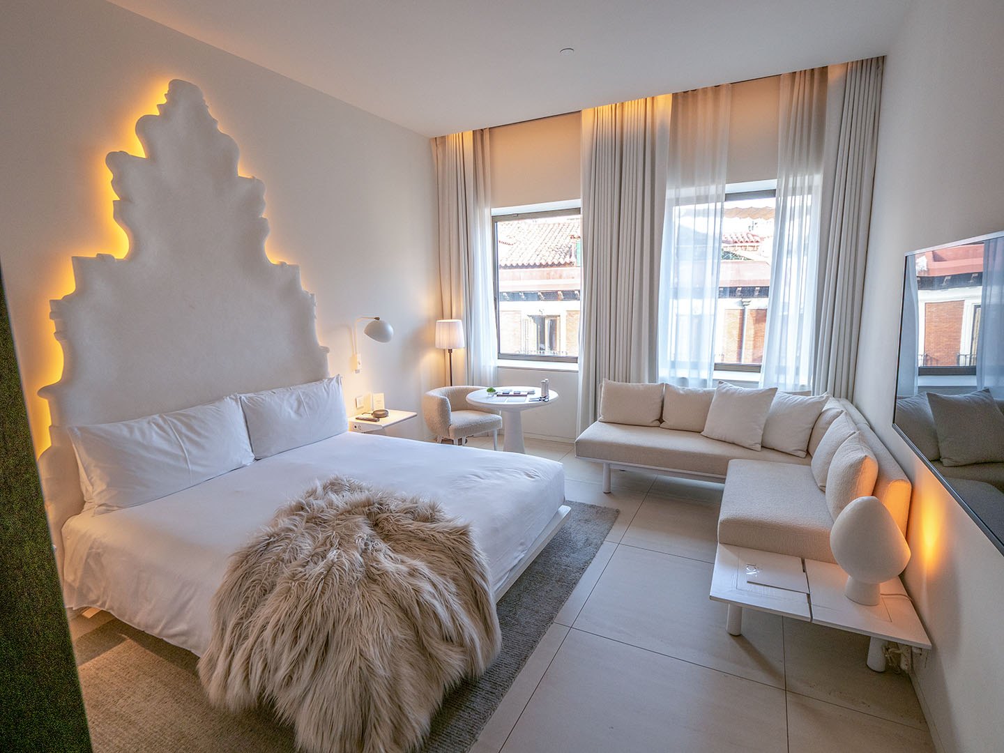 A guest room with a white sculpted, leaf-shaped headboard lit from behind, a fur throw on the bed and a white sofa by the windows.