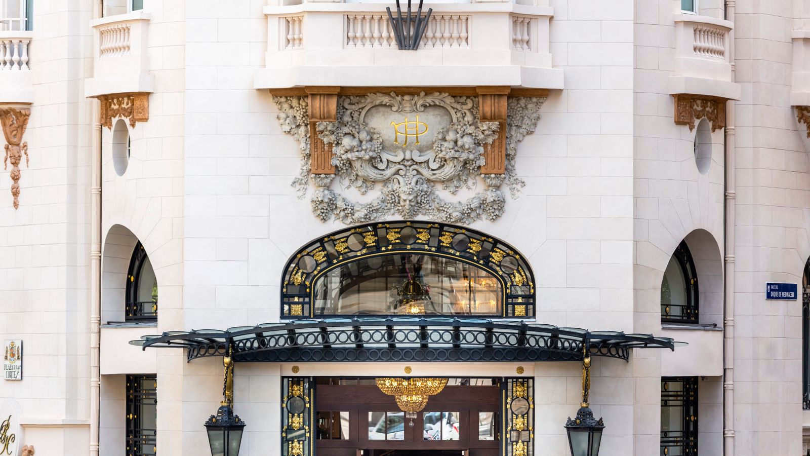 The main entrance: a glass and wrought-iron canopy below a carved stone cartouche with a gilded monogram, with tile plaques for Plaza de las Cortes and Calle del Duque de Medinaceli on the facade.