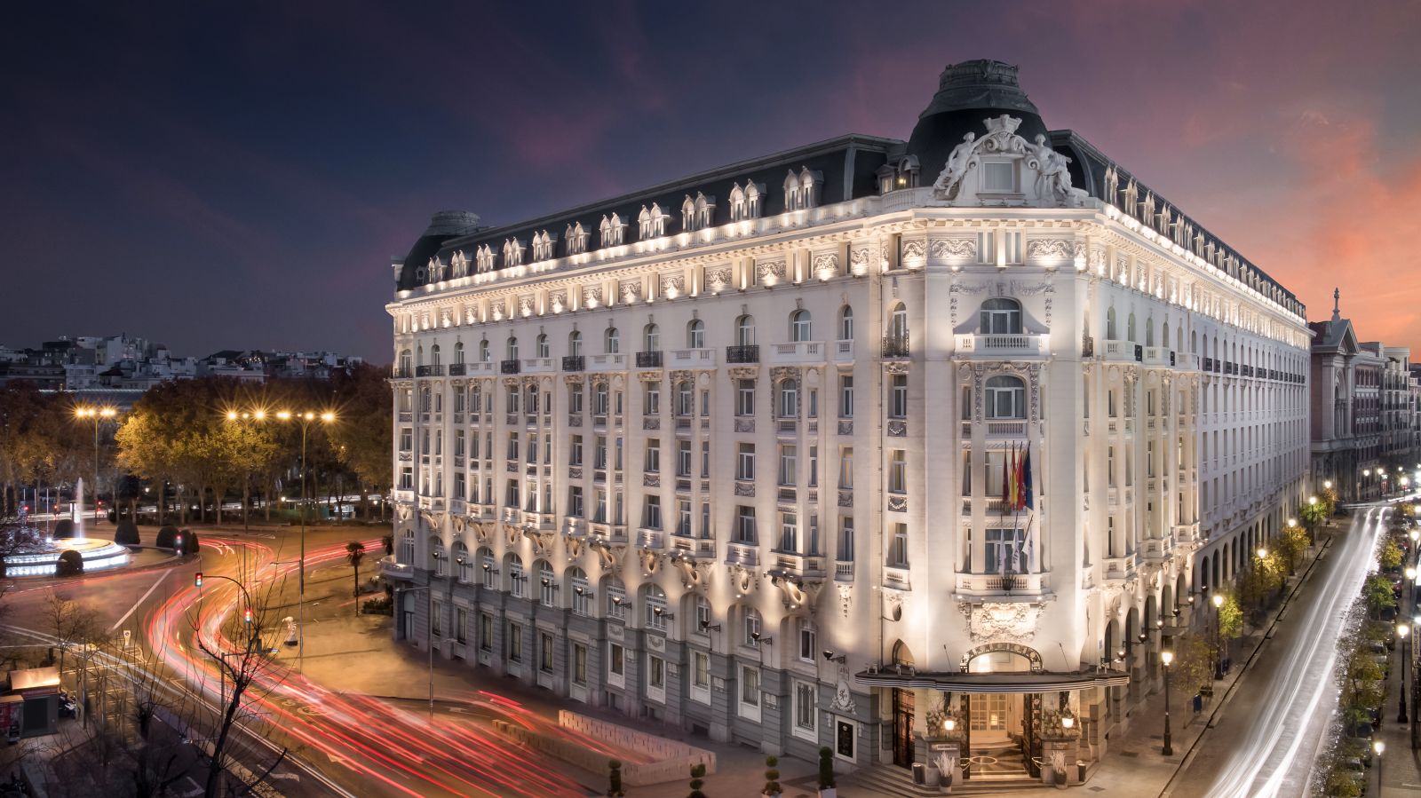 The hotel's full white Belle Epoque block at dusk, its facade floodlit under a domed corner tower, with traffic light-trails and a fountain roundabout on the near side.