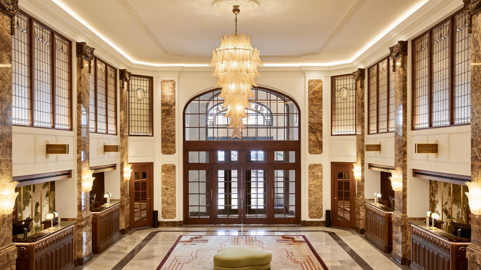 The entrance hall seen from above: a tiered glass chandelier over a rug and green ottoman, marble-clad pillars, leaded-glass panels and reception desks along both sides.