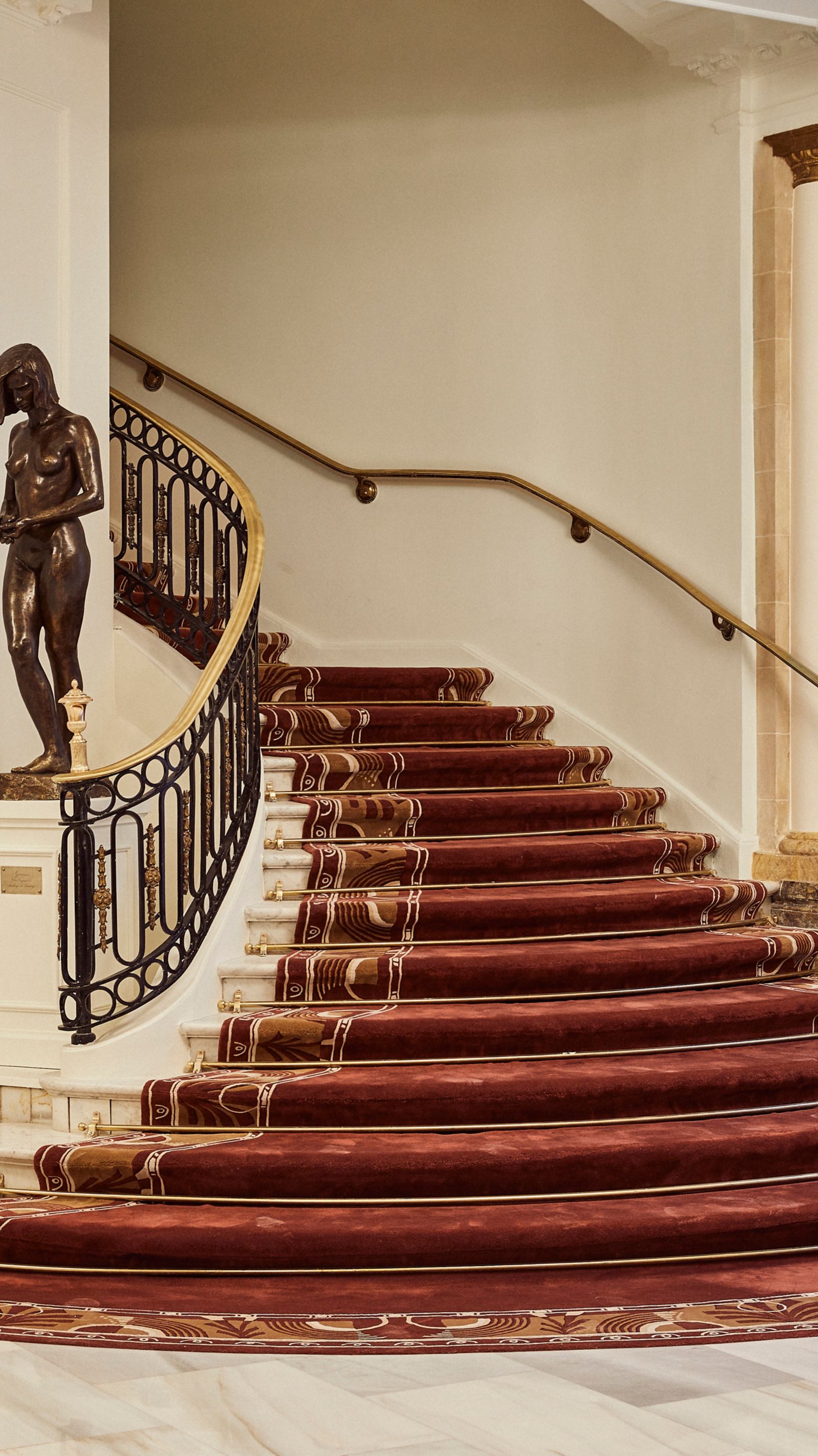 The curved main staircase with a red patterned runner held by brass stair rods, a brass handrail on a wrought-iron balustrade and a bronze figure on a marble pedestal.