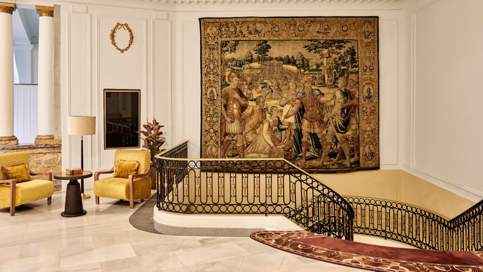 A stair-hall landing with a large classical tapestry on the wall, a wrought-iron and brass balustrade, marble floor and two yellow velvet armchairs.