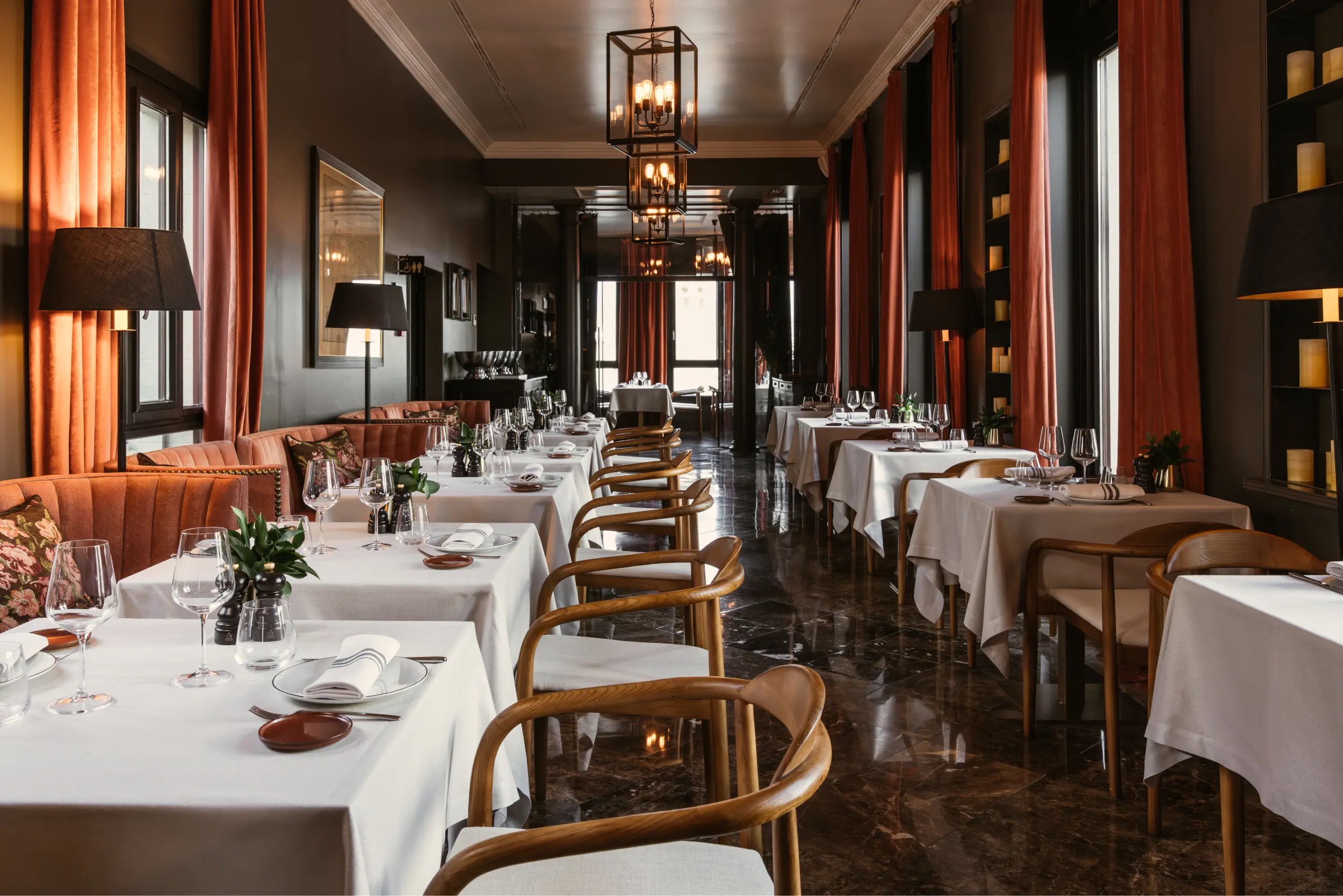 A long dining room with white-clothed tables, rust-orange banquettes and floor-length curtains, black lantern pendants and a polished dark marble floor.