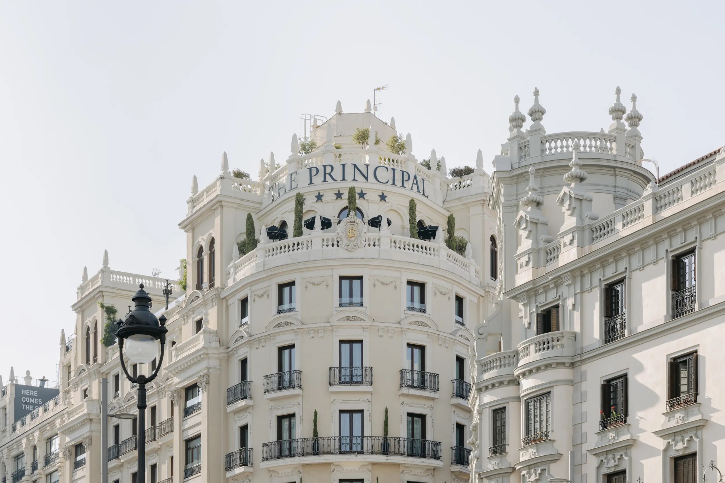 The hotel's curved corner facade topped by 'The Principal' lettering, with a balustraded rooftop terrace, cypresses and rows of wrought-iron balconies.