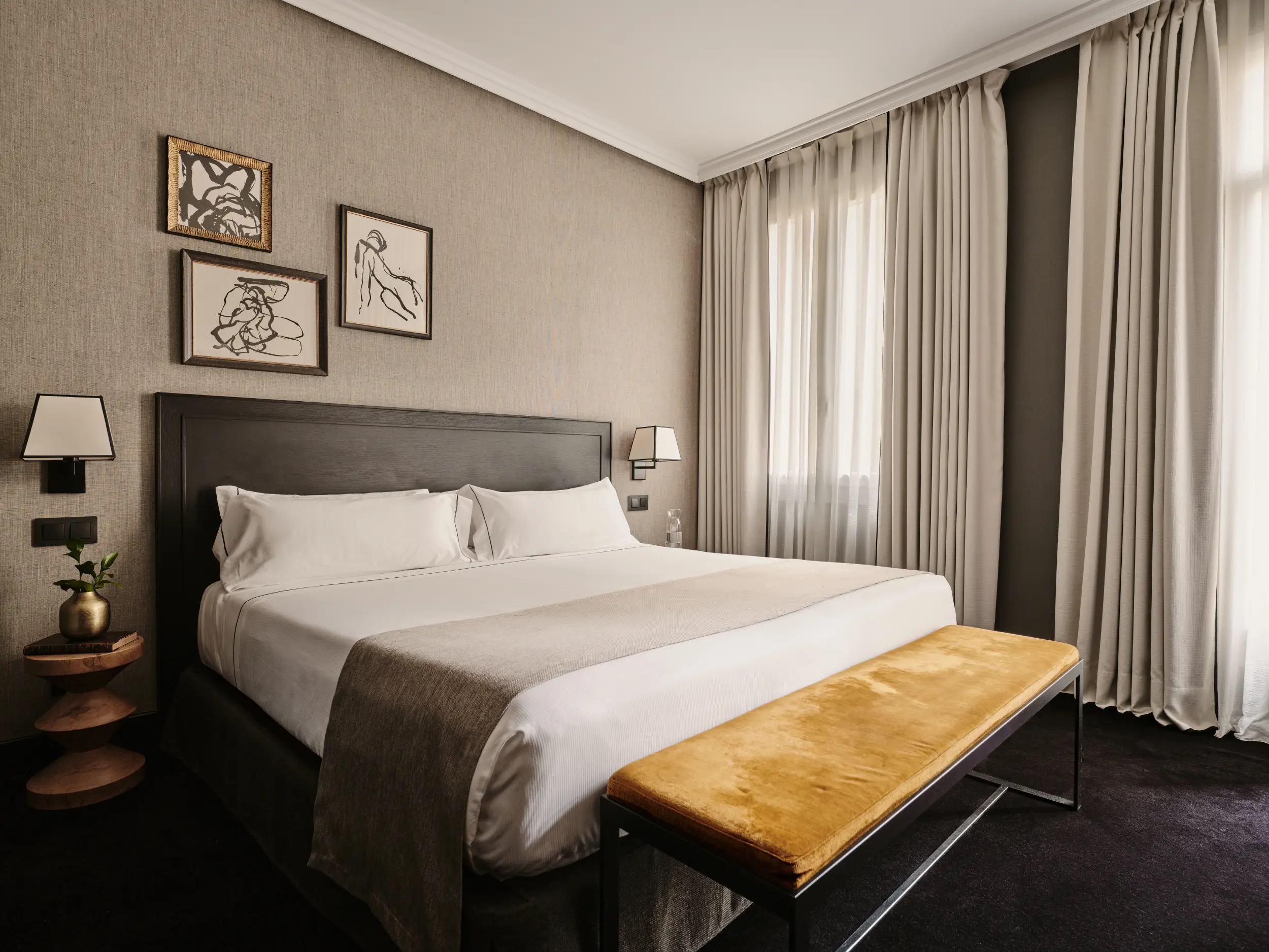 A guest room with three framed ink sketches above a dark wooden headboard, linen-textured walls and a mustard velvet bench at the foot of the bed.