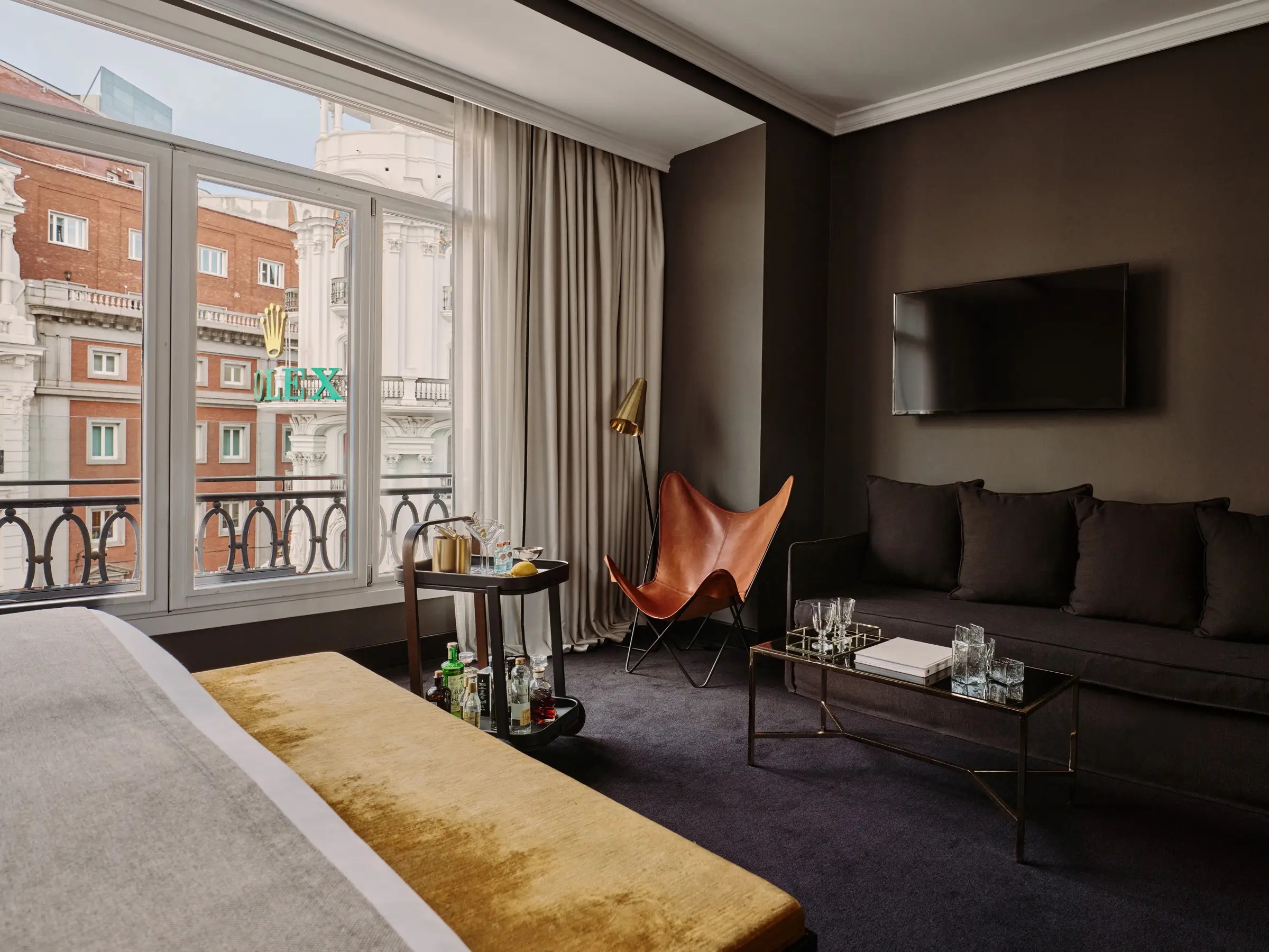 A room with a bed and mustard bench in the foreground, a drinks trolley, a leather butterfly chair and a charcoal sofa beneath a wall-mounted TV, with neighbouring facades through the window.