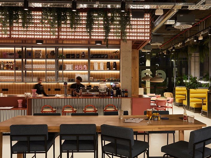 Bar and cafe at The Social Hub Barcelona Poblenou with pink tiled counter, backlit shelving, hanging plants, and mustard-yellow booth seating